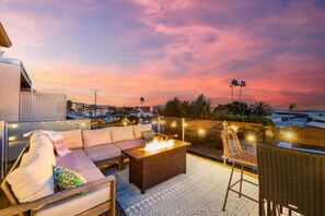 Beautifully arranged outdoor balcony at twilight, featuring a luxurious lounge area with a sectional sofa adorned with colorful cushions, a fire table emitting a warm glow, and stylish string lights that add a charming ambiance.