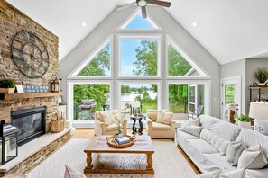 A bright and spacious lakefront living area with large floor-to-ceiling windows, cozy seating, a stone fireplace, and open access to the kitchen and outdoor deck.