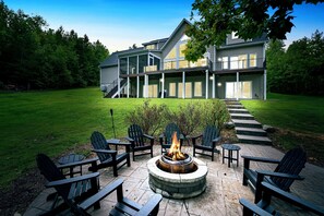 A spacious backyard with a fire pit seating area overlooking the lake, surrounded by greenery and showcasing a stunning multi-level lake house with large windows and outdoor spaces.
