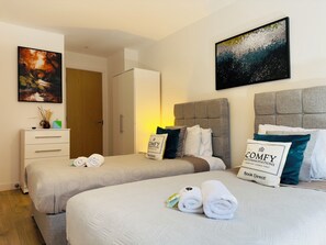 A modern twin bedroom with two comfortable single beds, plush headboards, and stylish decor. The room features a wardrobe for storage, a chest of drawers, and elegant wall art. Soft bedding and accent pillows create a cozy feel, making it a perfect space for relaxation