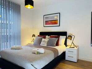 This cozy bedroom by Comfy Accommodations features a plush double bed, soft pillows, fresh towels, and a stylish grey throw. Modern bedside lamps and abstract wall art create a relaxing ambiance, with natural light from floor-to-ceiling blinds