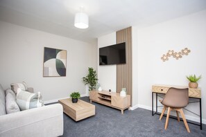A cozy living room featuring a modern TV stand, a comfortable sofa, a coffee table, and a desk area with a stylish chair and plants.