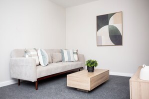 A minimalist living area with a beige sofa adorned with light blue pillows, a simple wooden coffee table with a small potted plant, and a geometric wall art piece.
