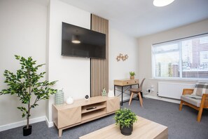 A cozy living room with a wooden TV stand, house plants, a minimalist coffee table, and a desk area with a chair. The space is well-lit with natural light from a window, adding a relaxed ambiance.