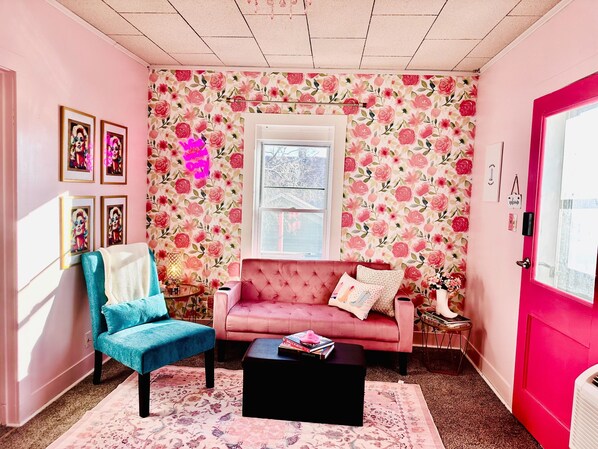 This uniquely decorated room features vibrant pink walls complemented by a bold floral wallpaper and a plush pink velvet loveseat, creating a fun and inviting space. A striking teal armchair and eclectic artwork add pops of color and personality to this cozy and stylish retreat.