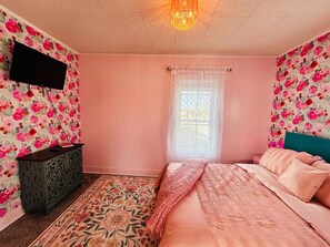 This charming bedroom features soft pink walls accented by vibrant floral wallpaper on select walls, creating a cozy and cheerful ambiance. A plush bed with pink linens and a teal headboard invites relaxation, while a decorative rug and a unique cabinet add character to the space.