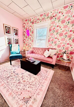 This cozy living room offers a playful ambiance with its soft pink walls, a comfortable pink velvet sofa, and a bold floral wallpaper as a focal point. The eclectic mix of artwork, a vibrant teal chair, and a patterned rug contributes to its unique and inviting feel.