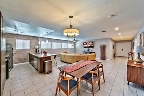 The open-plan living area features a dining space with a wooden table, six blue-cushioned chairs, and a chandelier. A curved beige sectional faces a wall-mounted 65" Roku TV. The kitchen with an island and pendant lights is also in view.