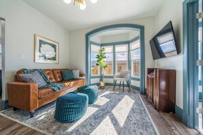  Inviting living space with natural light streaming through bay windows. Decorated with a comfortable leather sofa, two plush ottomans, and a chic armchair. Modern decor with plants and framed art, enhancing the cozy feel.