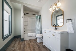 Modern bathroom with a clean, bright aesthetic. Features a bathtub with a stylish teal shower curtain, a white vanity with gold accents, and a large mirror with globe lights. Plenty of natural light from the window. Cozy and functional.