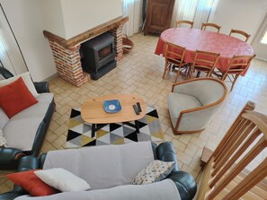 Vue d'ensemble du sĂ©jour avec les deux canapĂ©s, un fauteuil, le poĂȘle Ă bois ainsi qu"une table ovale avec ses six chaises. PiĂšce situĂ©e Ă l'est.