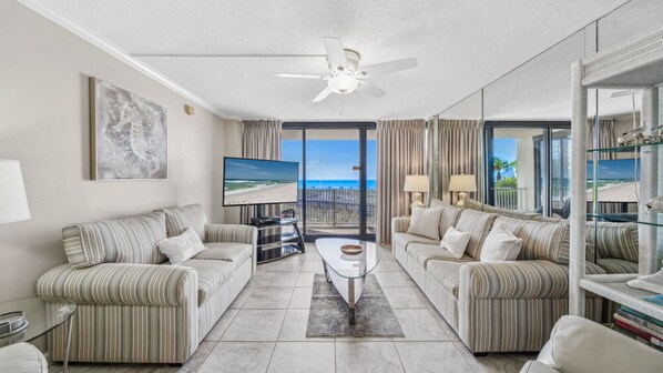 Relax in a bright, beachy living room with cozy striped sofas, a glass coffee table, and stunning Gulf views. Floor-to-ceiling doors open to a private balcony, with tile floors and coastal decor completing the space.