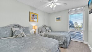 This charming guest bedroom offers two full-size beds adorned with calming seafoam bedding and subtle shell patterns, creating a peaceful retreat after a day in the sun. Natural light fills the space through the large window, which frames a tranquil view 