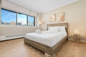 A serene and sunlit bedroom with a cozy bed, warm wood tones, and stunning city views