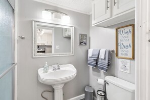 A fresh and inviting bathroom with a pedestal sink, soft towels, and bright lighting. Thoughtful touches like extra storage and a decorative sign add to the charm.