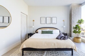 Bedroom with a black cane headboard, white linens, and a beige textured blanket. Features matching black nightstands, gold wall sconces, and abstract wall art. A large round mirror, woven basket planter with a fiddle-leaf fig, and floor-to-ceiling windows