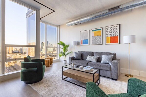 Spacious living area with a gray fabric sofa, two plush green swivel chairs, 65" TV, media stand, and a glass-top coffee table with wood accents. Large windows with view of One World Trade Center, complemented by modern artwork, gold-accented floor lamps,