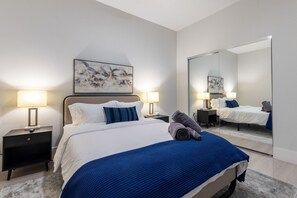 Cozy queen bedroom with a white bed, blue accents, and mirrored sliding closet doors. The room has modern bedside tables, soft lighting, 55 " Smart TV, full length closet sliding mirrors and a neutral-toned rug on light wood flooring. Foldable working des