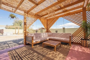 This outdoor seating area under a wooden pergola features a cozy L-shaped sofa with beige cushions, a matching coffee table, and hanging plants. Perfect for relaxing or social gatherings!