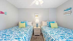 This cheerful coastal-themed bedroom features two twin beds with vibrant ocean-print bedding and playful accent pillows. Nautical wall art and a wicker nightstand with a beachy lamp complete the inviting, family-friendly space.