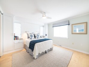 This cozy bedroom is bathed in natural light, showcasing chic bedding, wooden flooring, and tasteful decor that invites relaxation and comfort.