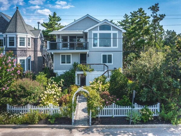 This charming coastal home features lush landscaping, a welcoming front entry, and an inviting upper deck perfect for enjoying ocean views.