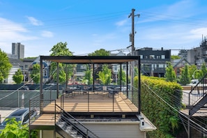 The property rooftop terrace with a modern metal pergola, wooden decking, a lounge seating area with black chairs and a coffee table, a metal staircase leading up from the ground level, and a lush green wall bordering the terrace.