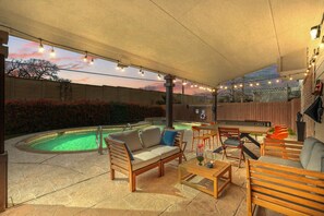 This covered backyard patio is fully furnished with a conversation set, an outdoor dinner table that doubles for table tennis, twinkling string lights, and a view of the pool.