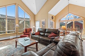 Spacious living area with vaulted ceilings, large windows providing ample sunlight, and electric reclining sofas surrounding the cozy fireplace.