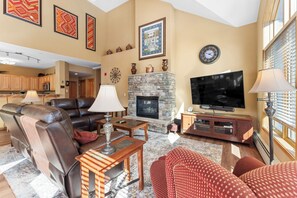 Spacious living area with vaulted ceilings, large windows providing ample sunlight, and electric reclining sofas surrounding the cozy fireplace.