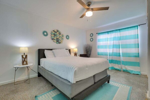 Relax in this tranquil bedroom with coastal-inspired decor, a plush queen bed, soft lighting, and vibrant aqua-striped curtains that add a refreshing touch.