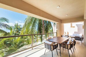 This image showcases a luxurious balcony with a breathtaking ocean view. The spacious outdoor area is elegantly furnished with comfortable seating and a dining table, perfect for relaxing or enjoying meals while surrounded by tropical greenery. 
