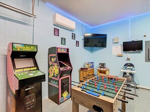 A pastel blue game room features arcade machines, a foosball table, and two flat-screen TVs. Small framed prints decorate the walls, and a white air conditioning unit is mounted above. Blue LED strip lighting runs along the ceiling, while wooden storage chests hold board games. A high chair sits nearby.