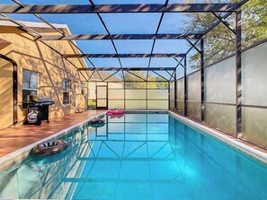 A covered patio houses a turquoise rectangular pool reflecting the surroundings. Dark pool floats rest at the edge, while a charcoal grill sits nearby. Beige walls and a metal-framed translucent roof provide shade, with glimpses of trees and sky beyond.
