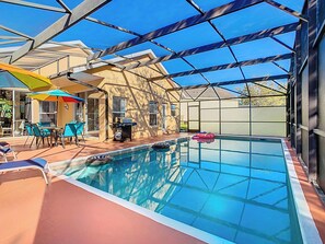 A screened-in pool area with clear turquoise water, surrounded by a light terracotta deck. A small inflatable float drifts in the shallow end. Colorful umbrellas shade a patio with a teal dining set. A beige house and a charcoal grill sit in the background. Lounge chairs line the deck, while the metal-framed enclosure shelters the space under a bright blue sky.