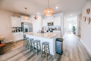 Step into this spacious, bright, and beautifully designed kitchen, the heart of the home! Featuring a large island with seating for four, stainless steel appliances, and elegant white cabinetry, this space is perfect for cooking, dining, and making memories.
Warm coastal touches, including woven pendant lighting and nautical decor, create a relaxing and inviting atmosphere. Th...