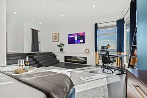 This modern bedroom boasts a cozy fireplace, sleek black steps, and a mounted smart TV. Floor-to-ceiling windows showcase stunning ocean views, complemented by navy curtains, a stylish workspace, and a plush bed.