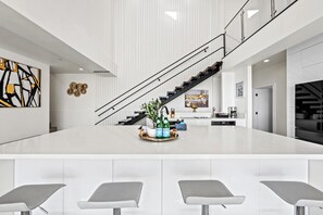 This open-concept space features a sleek white kitchen with a spacious island and chic gray barstools. A bold black staircase with glass railings leads upstairs, complemented by modern artwork and elegant decor for a stylish ambiance.