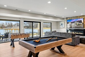 Game Room: Spacious entertainment area featuring a pool table, foosball, comfortable seating, cozy fireplace, and panoramic lake views, perfect for family fun and friendly competition.