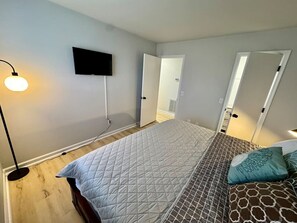 Cozy bedroom with a king bed, soft lighting, nightstands, a mounted TV, and natural light.