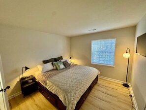 Cozy bedroom with a king bed, soft lighting, nightstands, a mounted TV, and natural light.