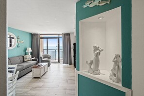 A mermaid-themed beach cove condo with a full couch, queen sofa sleeper, and elegant mermaid statues. Soft coastal tones complement ocean views through large glass doors leading to the balcony, filling the airy space with seaside charm.