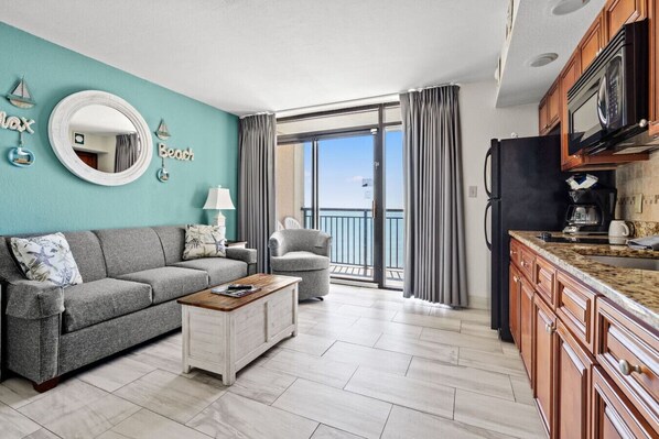 A bright beach cove 1-bedroom condo living room with a full couch and queen sofa sleeper, decorated in a coastal theme. Large glass doors open to a balcony with stunning ocean views, letting in natural light and a refreshing sea breeze.