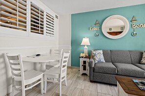 A bright living and dining area with a clean white table and cozy seating. The open layout feels airy, with natural light enhancing the coastal decor. Soft tones and ocean views create a relaxing, inviting space perfect for dining and lounging.