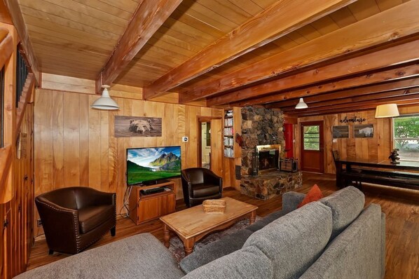 Gather and relax in our warm living room, featuring a gray sectional sofa, leather armchairs, a stone fireplace, and a scenic dining area—all bathed in natural light