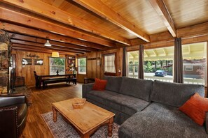 Gather and relax in our warm living room, featuring a gray sectional sofa, leather armchairs, a stone fireplace, and a scenic dining area—all bathed in natural light