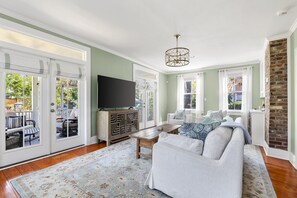 This bright and inviting living room seamlessly connects to the outdoor space through French doors, allowing for an effortless indoor-outdoor lifestyle. The soft green walls, natural light, and cozy furnishings create a welcoming ambiance.