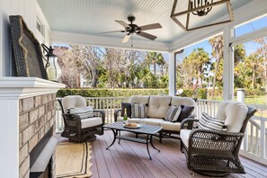 This covered porch lounge is a serene retreat with comfortable wicker seating, plush cushions, and a ceiling fan for added relaxation.