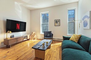 Unwind in this stylish and sunlit living room, featuring a plush emerald green sofa, sleek modern décor, and a large flat-screen TV for ultimate relaxation.