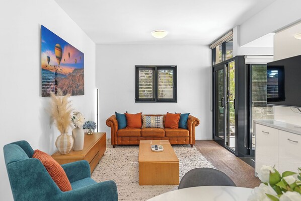 Unwind in our sun-drenched living room, where a plush teal armchair invites you to relax and a warm, inviting orange sofa beckons for cozy evenings.
